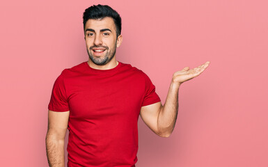 Wall Mural - Hispanic man with beard wearing casual red t shirt smiling cheerful presenting and pointing with palm of hand looking at the camera.