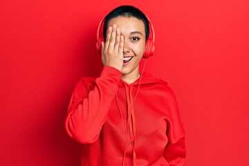 Beautiful hispanic woman with short hair listening to music using headphones covering one eye with hand, confident smile on face and surprise emotion.