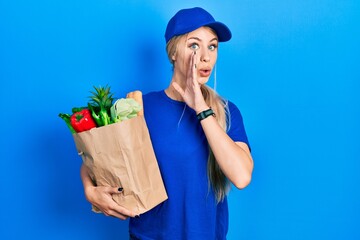 Poster - Young caucasian woman wearing courier uniform with groceries from supermarket hand on mouth telling secret rumor, whispering malicious talk conversation