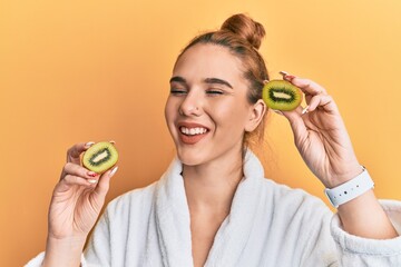 Poster - Young blonde woman wearing bathrobe holding fresh kiwi smiling and laughing hard out loud because funny crazy joke.