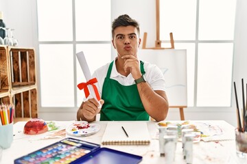 Wall Mural - Young hispanic man at art studio holding degree looking at the camera blowing a kiss on air being lovely and sexy. love expression.