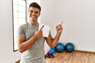 Poster - Young hispanic man wearing sportswear and towel at the gym smiling and looking at the camera pointing with two hands and fingers to the side.