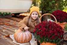 Happy Little Girl Sitting On Porch Of House With Chrysanthemums Potted And Pumpkins. Home Fall Decoration For Halloween Or Thanksgiving. Smilling Child In Autumn Garden With Yellow Pumpkins And Flower