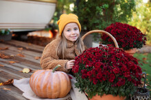 Happy Little Girl Sitting On Porch Of House With Chrysanthemums Potted And Pumpkins. Home Fall Decoration For Halloween Or Thanksgiving. Smilling Child In Autumn Garden With Yellow Pumpkins And Flower