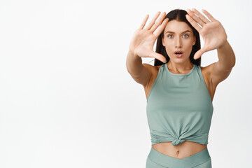 Wall Mural - Image of sportswoman looking through hand frames with surprised, fascinated face expression, standing in sports clothing over white background