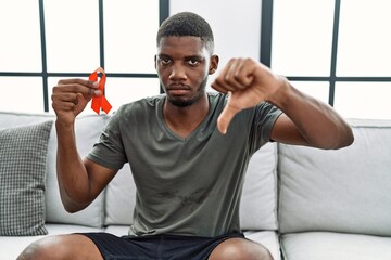 Young african american man holding support red ribbon sitting on the sofa with angry face, negative sign showing dislike with thumbs down, rejection concept