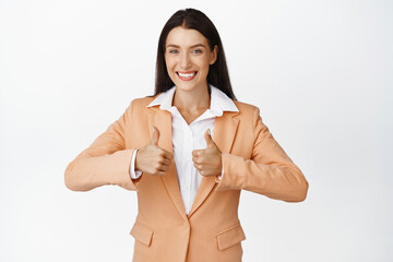 Wall Mural - Smiling businesswoman showing thumbs up, approve smth good. Saleswoman give positive feedback, standing in suit against white background