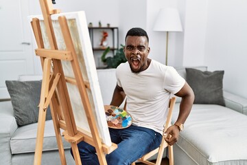 Wall Mural - Young african man painting on canvas at home angry and mad screaming frustrated and furious, shouting with anger. rage and aggressive concept.