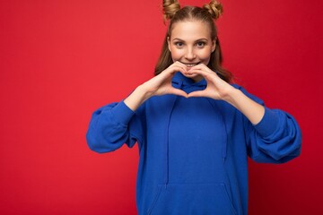 Wall Mural - Young happy positive attractive blonde woman with two horns with sincere emotions wearing stylish bright blue hoodie isolated on red background with copy space and showing heart gesture