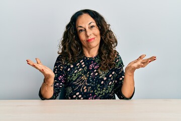 Poster - Beautiful middle age woman wearing casual clothes sitting on the table clueless and confused expression with arms and hands raised. doubt concept.