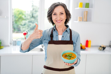 Wall Mural - Photo of age funny lady hold soup show thumb up wear apron jeans shirt at kitchen alone