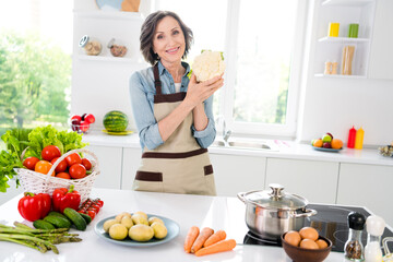 Photo of cheerful positive charming old woman hold hands cauliflower good mood indoors inside house home kitchen