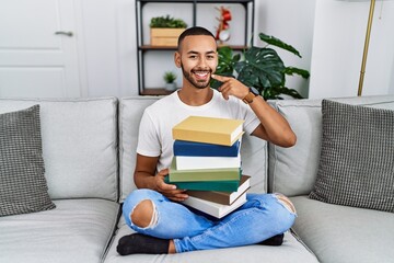 Canvas Print - African american young man holding a pile of books sitting on the sofa smiling cheerful showing and pointing with fingers teeth and mouth. dental health concept.