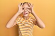 Beautiful brunette little girl wearing casual striped t shirt doing ok gesture like binoculars sticking tongue out, eyes looking through fingers. crazy expression.