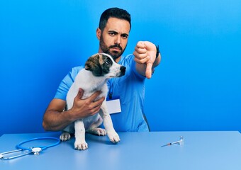 Canvas Print - Handsome hispanic veterinary man with beard checking dog health looking unhappy and angry showing rejection and negative with thumbs down gesture. bad expression.