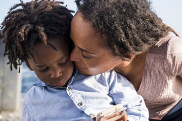 loving african american mother kissing her little son watching video or playing game using mobile ph