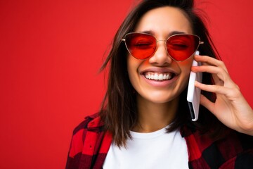 Closeup photo of attractive positive smiling young brunet female person wearing stylish red shirt white t-shirt and red sunglasses isolated over red background communicating on mobile phone with close