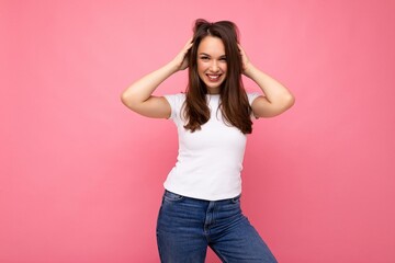 Wall Mural - Photo portrait of young beautiful smiling hipster brunette woman in white t-shirt with mockup. Sexy carefree female person posing isolated near pink wall with empty space in studio. Positive model