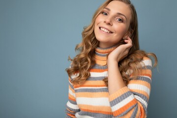 Wall Mural - Portrait of young winsome attractive happy smiling blonde woman with wavy-hair wearing striped sweater isolated over blue background with empty space