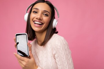 Wall Mural - Closeup photo of beautiful happy smiling young woman wearing stylish casual outfit isolated on background wall holding and showing mobile phone with empty display for mockup wearing white bluetooth