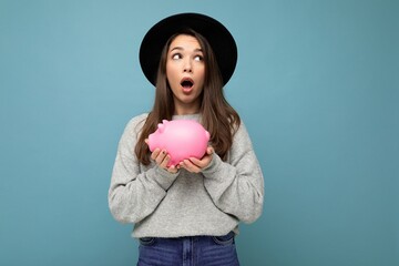 Wall Mural - Portrait of shocked amazed young pretty charming brunette woman wearing stylish gray sweater and black hat isolated over blue background with empty space and holding pink piggy box. Money box concept