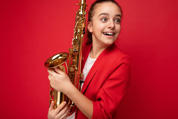 Photo shot of beautiful positive amazed surprised brunette female teenager wearing trendy red jacket standing isolated over red background wall holding saxophone looking to the side