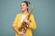 Shot of beautiful cute happy smiling brunette female teenager wearing trendy yellow jacket standing isolated over blue background wall holding saxophone looking to the side