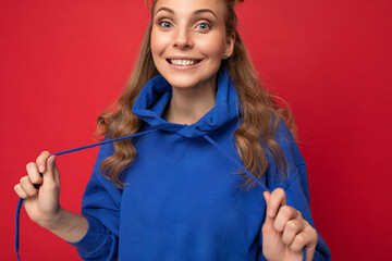 Wall Mural - Photo of young charming beautiful attractive happy smiling delightful blonde woman with sincere emotions wearing stylish hipster blue hoodie isolated over red background with copy space