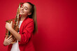 Beautiful cute positive smiling brunette little female teenager wearing trendy red jacket standing isolated over red background wall holding saxophone and dreaming with close eyes