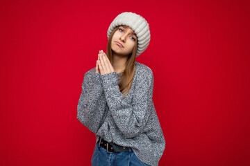 Wall Mural - Portrait of young pretty dark blonde woman with sincere emotions wearing grey sweater and beige knitted hat isolated over red background with free space and praying with pray gesture