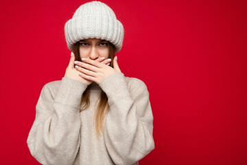 Wall Mural - Portrait of angry dissatisfied young beautiful dark blonde woman with sincere emotions wearing beige pullover and knitted hat isolated over red background with copy space and covers mouth with hands