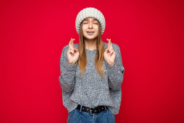 Wall Mural - Portrait of emotional young beautiful dark blond woman with sincere emotions wearing gray sweater and beige hat isolated over red background with empty space and gesturing fingers crossed with hope