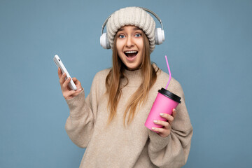 Portrait of positive happy amazing joyful young beautiful blonde woman with sincere emotions wearing beige pullover and knitted hat isolated over blue background with copy space and listening ti music