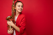 Side-profile Photo Shot Of Beautiful Positive Smiling Brunette Little Girl Wearing Trendy Red Jacket Standing Isolated Over Red Background Wall Holding Saxophone Looking To The Side