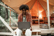 Young smiling African man with piercing sitting at glamping typing on laptop. Camping lifestyle. Low budget travel, holiday. Wi-fi connection information communication technology. Remote work