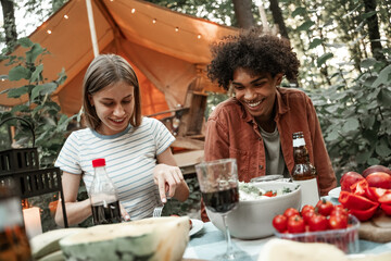 Wall Mural - Young multiracial couple having dinner at glamping, laughing after sunset. Happy millennials camping at open air picnic under bulb lights. Spending time with friends outdoors, barbeque party