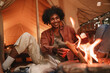© Marharyta Hanhalo - Hansome young curly african man roasting marshmallows on skewers over fire pit at campsite, enjoying outdoor glamping holiday with friends togetherness reopen after pandemic lockdown.