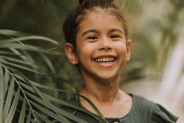 Wall Mural - The face of a little girl surrounded by tropical leaves. Closeup portrait of a beautiful swarthy baby with perfect skin and dark hair. Natural cosmetics, health, cleanliness, skin care, beauty concept