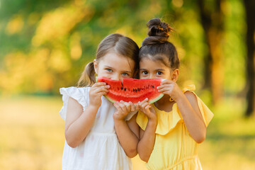 Two smiling girls are holding one slice of watermelon in their hands. Kids eat fruit outdoor. Healthy snack for children. Kids enjoying watermelon in the garden. Happy childhood. Space for text