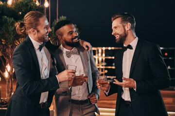 Wall Mural - Three handsome men in suits drinking whiskey and communicating while spending time on party