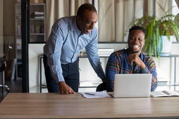 Happy Black office employees of different ages looking at laptop screen at workplace, laughing, having fun. African American coworkers reading email, making video call, watching presentation