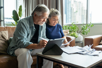 old retired asian senior couple checking and calculate financial billing together on sofa involved in financial paperwork, paying taxes online using e-banking laptop at living room home background