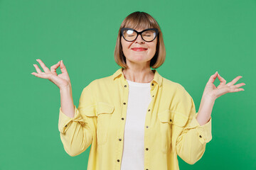 Wall Mural - Elderly happy woman 50s in glasses yellow shirt hold spreading hands in yoga om aum gesture relax meditate try to calm down isolated on plain green background studio portrait People lifestyle concept.