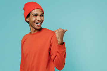 Young smiling happy fun african american man 20s in orange shirt hat point thumb finger back aside on workspace area copy space mock up isolated on plain pastel light blue background studio portrait