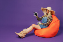 Full Body Young Woman With Halloween Makeup Mask In Straw Hat Scarecrow Costume Sit In Bag Chair Point Finger Aside Isolated On Plain Dark Purple Background Studio Celebration Holiday Party Concept.