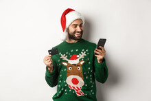 New Year, Online Shopping And Christmas Concept. Happy Bearded Man Buying In Internet With Credit Card And Phone, Wearing Santa Hat And Sweater, White Background