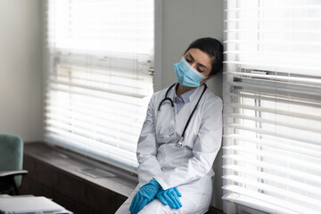 Tired female Indian doctor in protective medical mask and latex gloves leaning on wall, sleeping. Practitioner feeling exhausted and overworked after covid patients appointment on epidemic outbreak