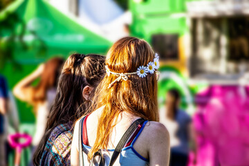 Two girls - one with red hair and daisy headband - wait in line at food truck at carnival - bright colors of pink and green