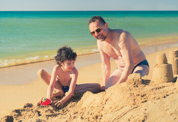 Bearded dad and little son are playing on the beach in the sand. Sea vacation on the shore on a sunny day.