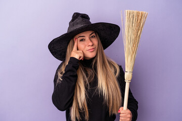 Wall Mural - Young Russian woman disguised as a witch holding a broom isolated on purple background pointing temple with finger, thinking, focused on a task.